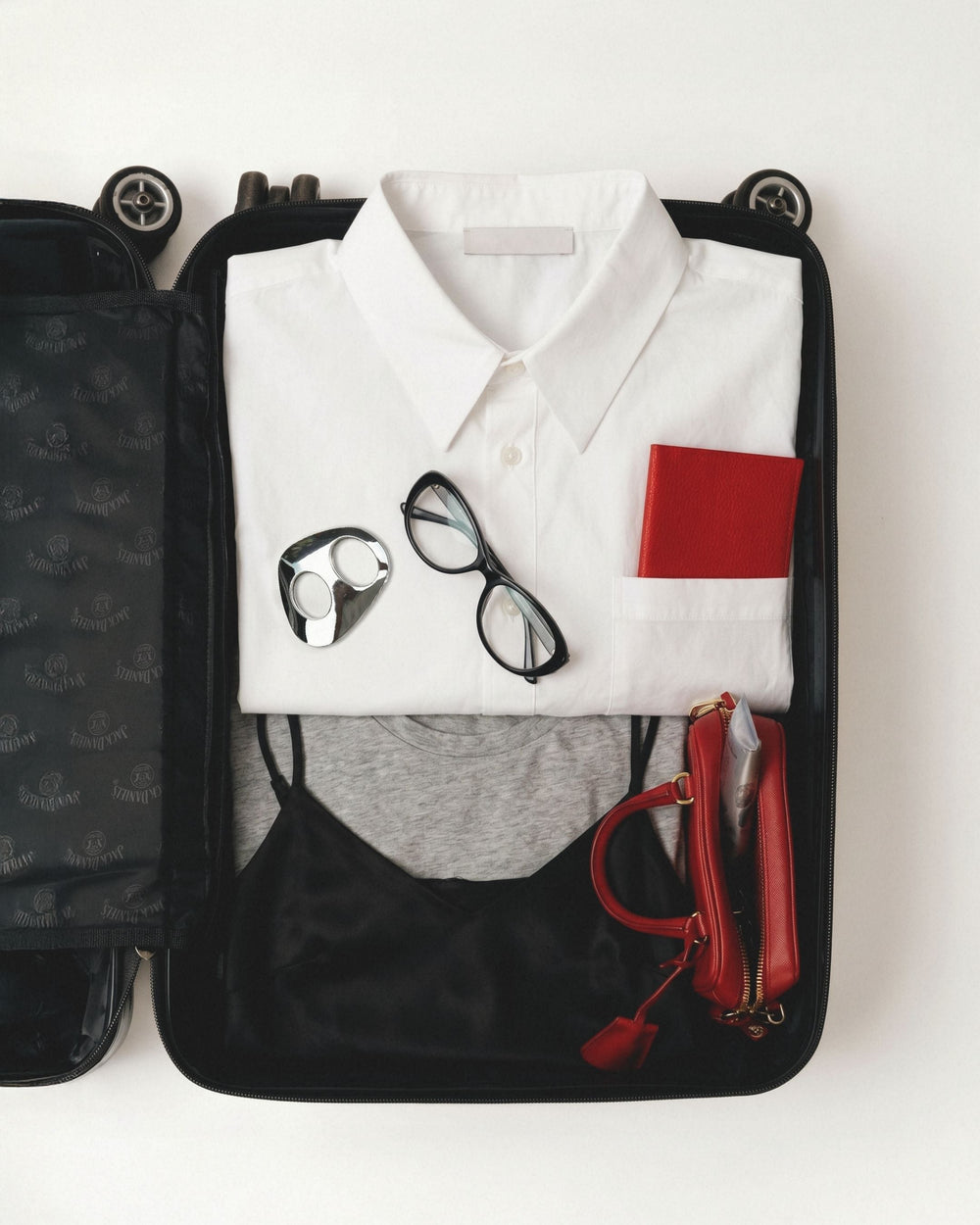 Open black suitcase flat-lay with white shirt, black glasses, red passport, and small red bag.