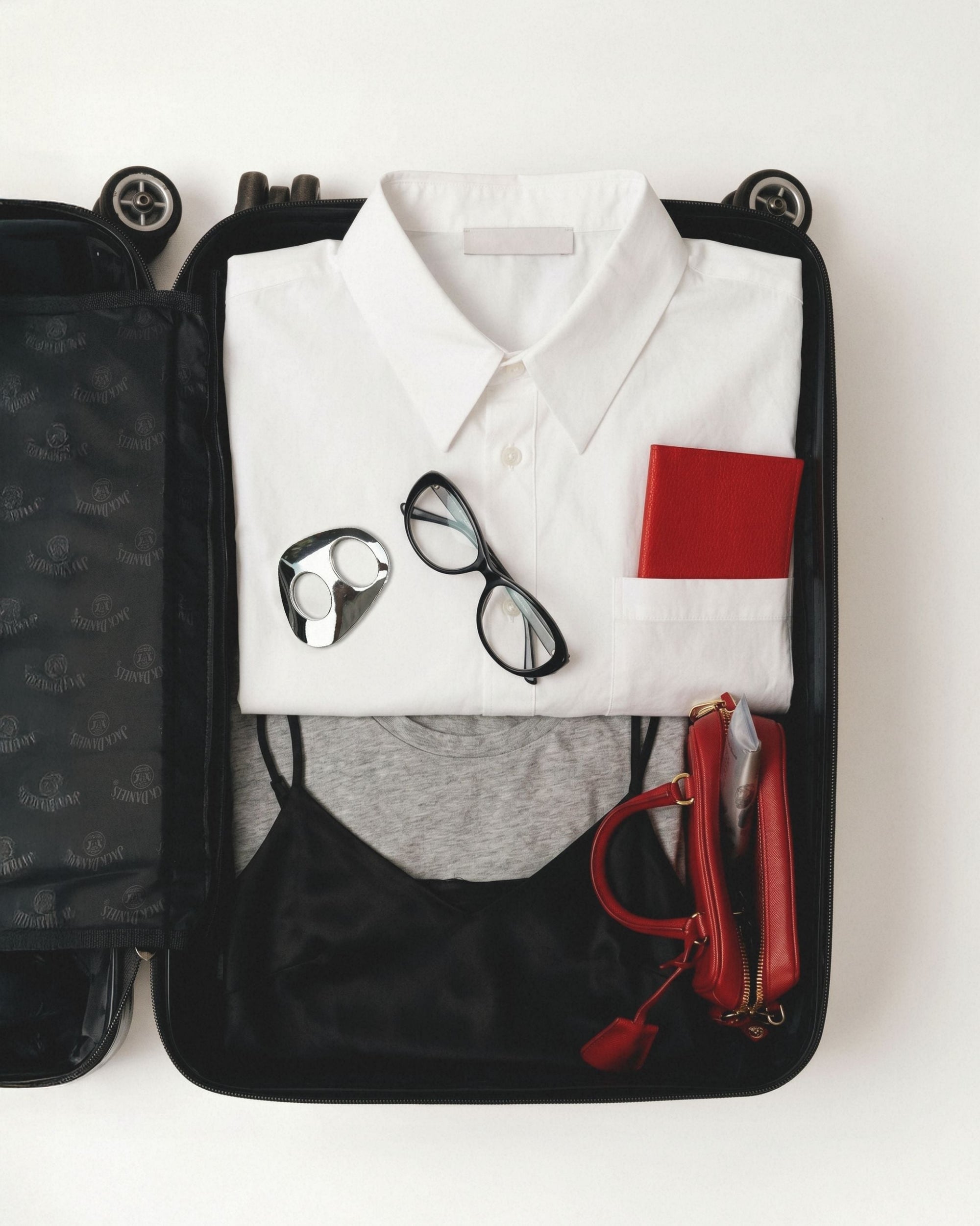 Open black suitcase flat-lay with white shirt, black glasses, red passport, and small red bag.
